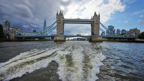 This urban landscape photograph was taken in the late afternoon during the summer season and prominently features the wake of a boat on the River Thames in London. The main subject of the image is the turbulent water in the foreground, leading the viewer’s eye towards the iconic Tower Bridge, which is an important piece of architecture in London. Tower Bridge stands tall over the river, with both its towers clearly visible and modern city buildings, such as the Shard, featured in the background. The scene showcases the blend of historic and contemporary architecture in London along the Thames, highlighting this well-known river crossing in the heart of the city. The photograph captures the dynamic movement of the river and the distinctive urban skyline behind Tower Bridge.